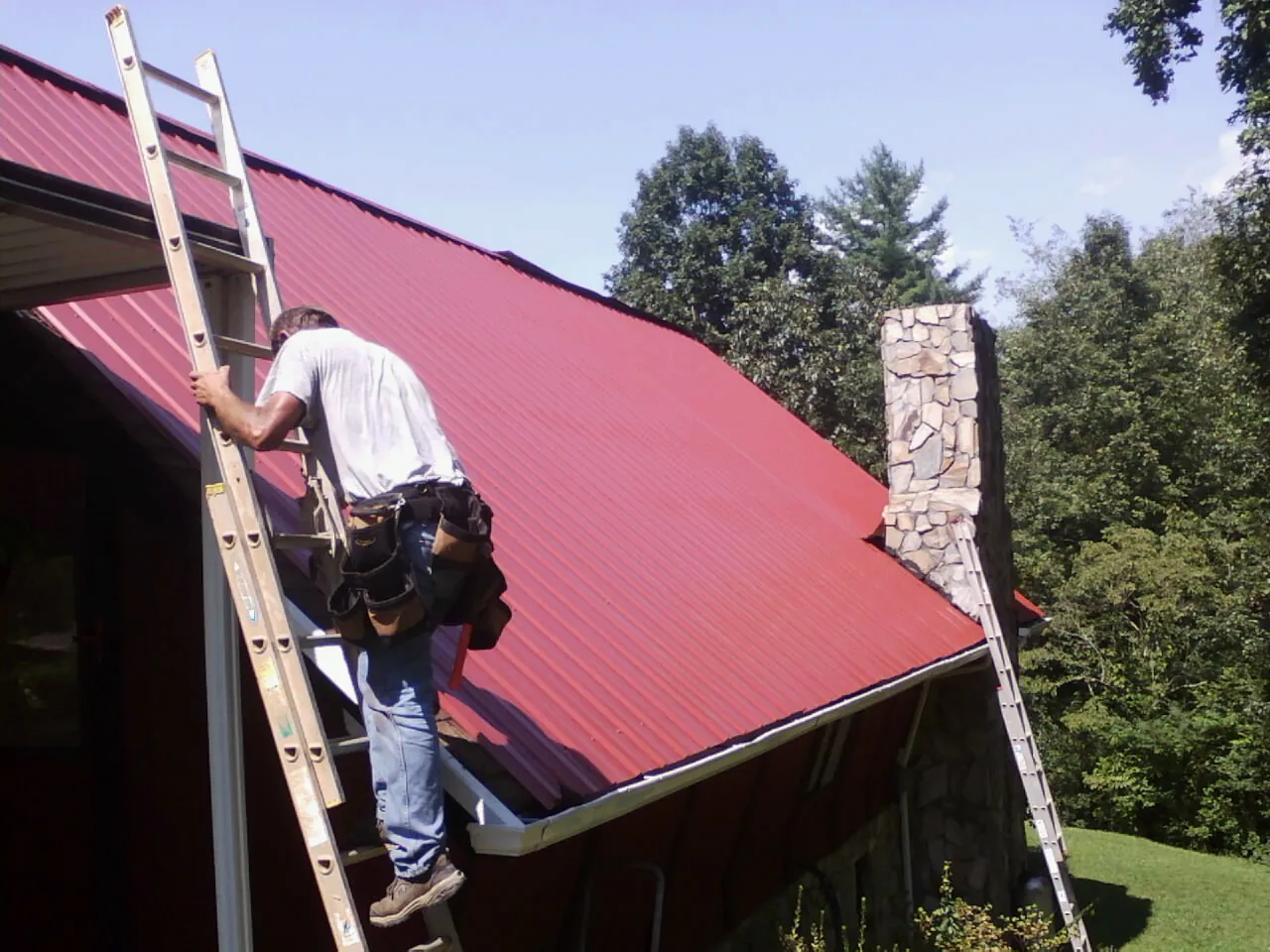 Roofer on a ladder beside a red metal roof and stone chimney — Soffit & Fascia Repair by Roofing Brothers in Sturbridge